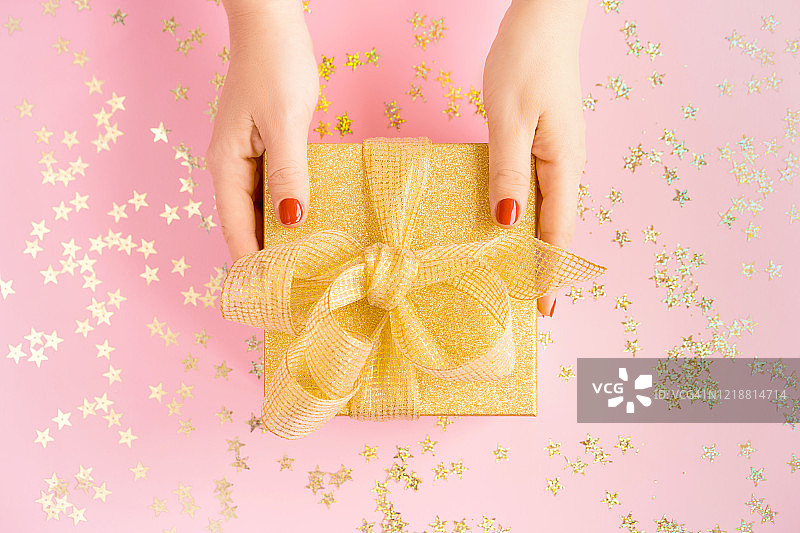 女人手持带黄色缎带的金色礼品盒，粉色背景图片素材