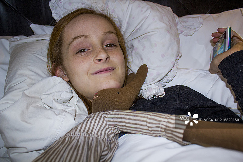 躺在床上，看着镜头，手里拿着手机，下巴底下放着毛绒兔子的红发短发褐发雀斑女孩特写图片素材