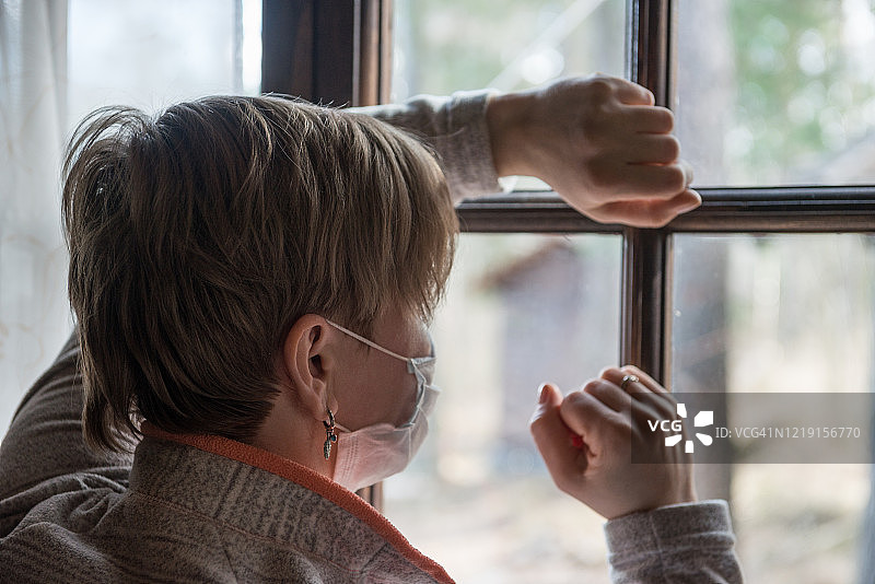 隔离期间悲伤的女人：戴着医用口罩的女孩望着窗外图片素材