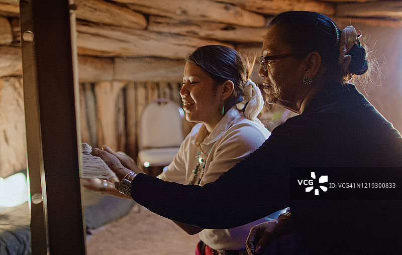 一位六十多岁的美国原住民（纳瓦霍族）祖母在霍根（纳瓦霍小屋）内，教她的十几岁孙女如何在织布机上编织图片素材