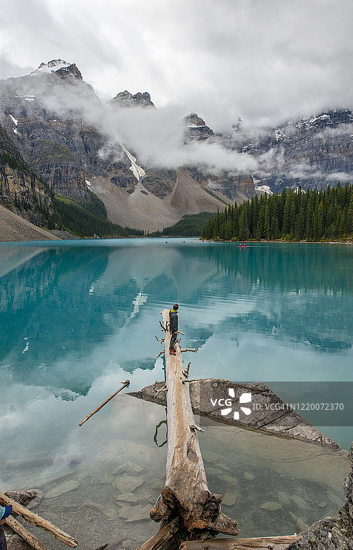 徒步男孩坐在梦莲湖边的树干上，眺望加拿大艾伯塔省班夫国家公园的落基山脉雪峰图片素材