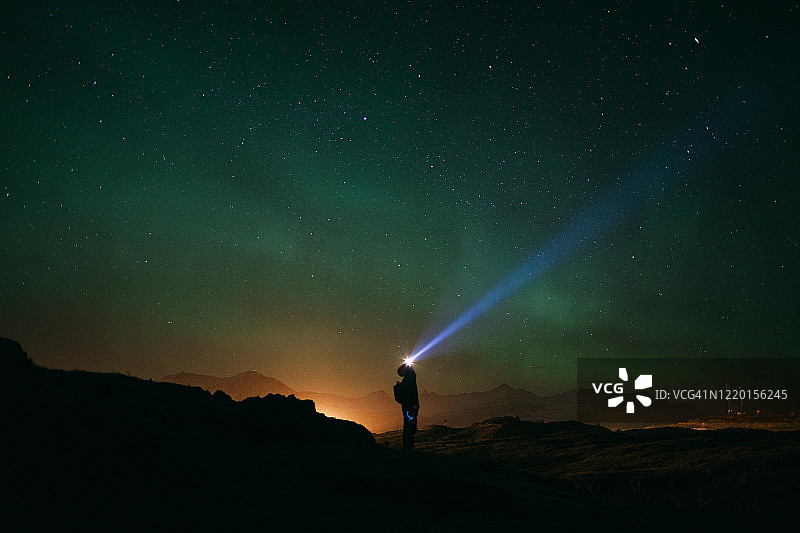 头戴手电筒的年轻男子站在绿色北极光下，光线照亮夜空图片素材
