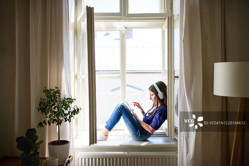 戴着耳机、手持平板电脑坐在窗台上的小女学生图片素材