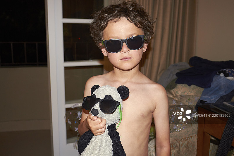 一个小男孩在家里抱着他的熊猫玩具，他们都戴着太阳镜看着镜头。男孩没穿衬衫，镜头从腰部以上拍摄图片素材