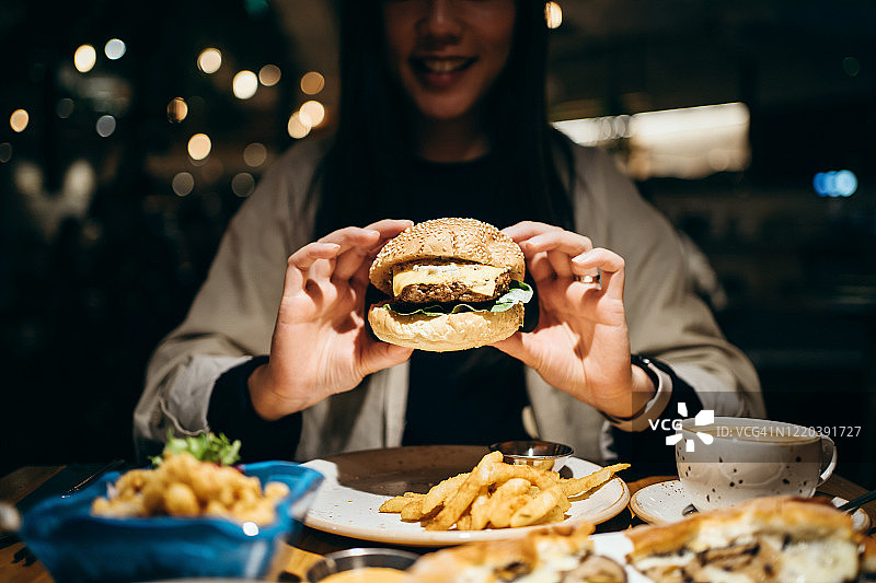 微笑的年轻女人在餐厅里享用美食，拿着汉堡准备咬一口图片素材