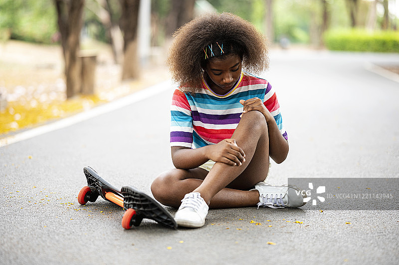 在滑板公园道路上受伤的滑冰者。非洲女孩在滑板事故后坐在路边图片素材