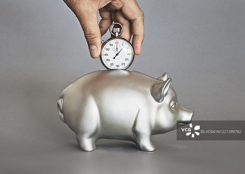 手持秒表在小猪储蓄罐上方图片素材