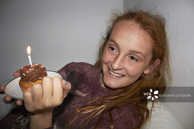 红发女孩手捧点燃生日蜡烛的纸杯蛋糕，笑对镜头图片素材