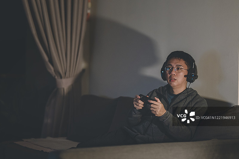 中国中年男子晚上独自在家玩电子游戏，戴着耳机，坐在沙发上图片素材