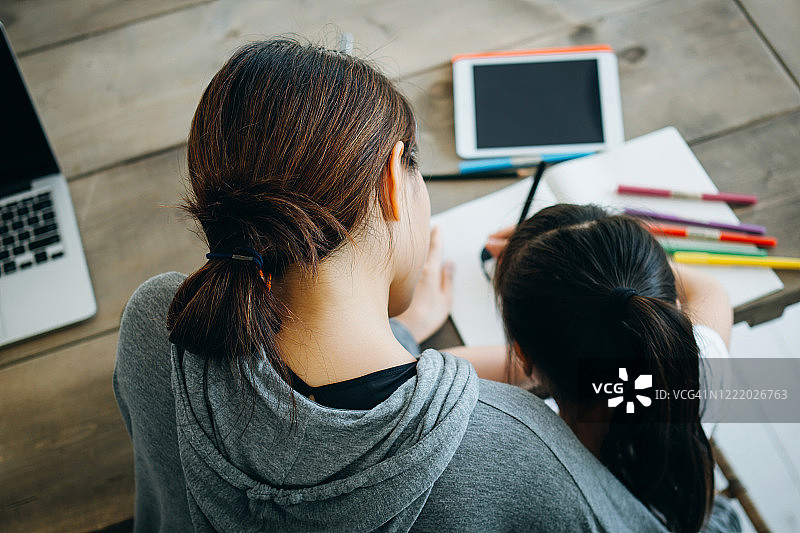 年轻的亚洲母亲在家用笔记本电脑工作，辅导女儿在家自学俯视图图片素材