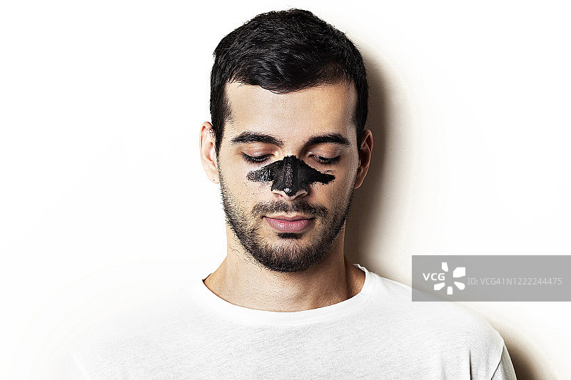 年轻男子脸上带着黑色木炭面膜，白色背景，年轻漂亮的男子脸上带着黑色撕脱面膜图片素材
