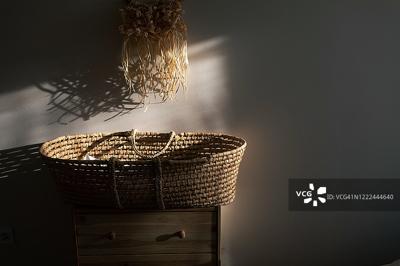 时尚斯堪的纳维亚新生婴儿房，柳条摇篮置于木制梳妆台上，墙上挂着拉菲亚流苏花边。米色背景墙的现代室内设计，木制家具和日落光线下手工制作的流苏花边。期待婴儿概念。图片素材