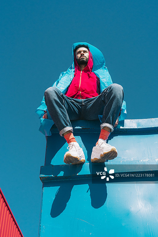 年轻男子身穿红色连帽夹克坐在蓝色天空下的集装箱上图片素材