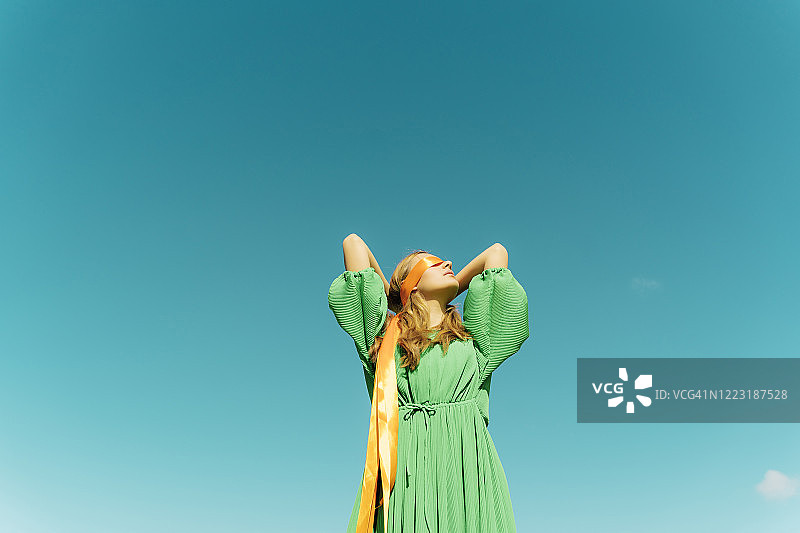 蓝天下，蒙着眼睛的年轻女人穿着绿色连衣裙图片素材