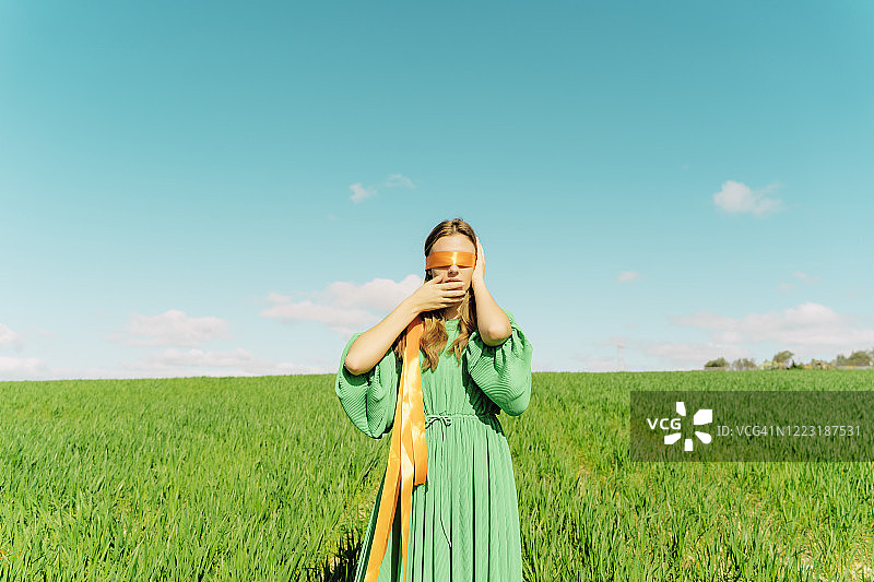 蒙着眼睛，穿着绿色连衣裙站在田野里的年轻女人图片素材