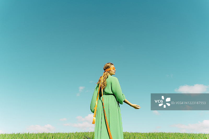 蒙着眼睛，穿着绿色连衣裙站在田野里的年轻女人图片素材