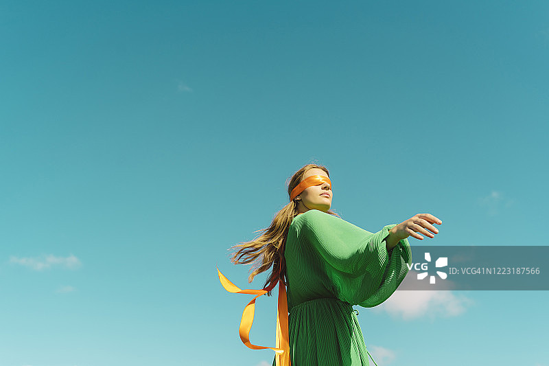 蓝天下，蒙着眼睛的年轻女人穿着绿色连衣裙图片素材
