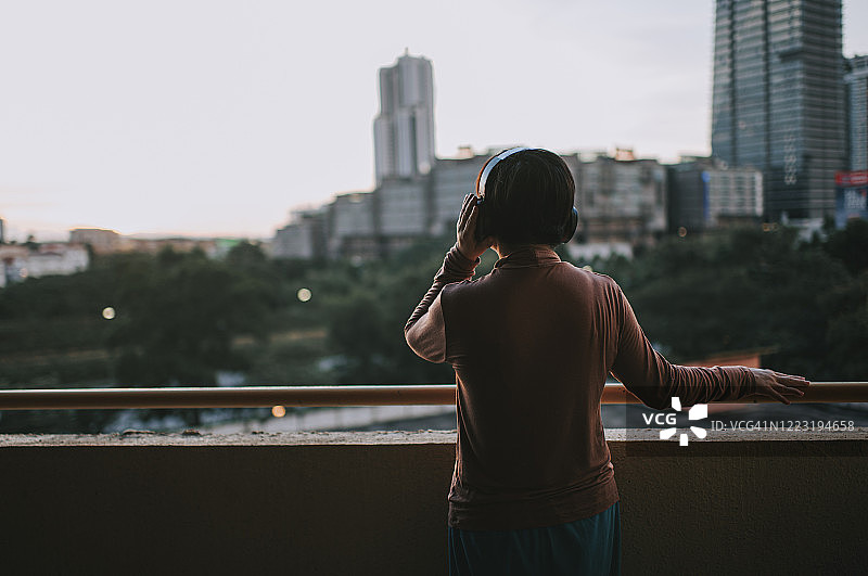 华人中年妇女在日落时分站在阳台上戴着无线耳机听音乐图片素材