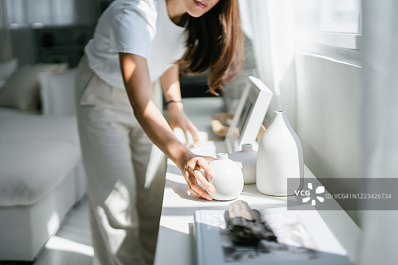 年轻亚洲女性在家里装饰和整理窗边架子上的相框和花瓶图片素材
