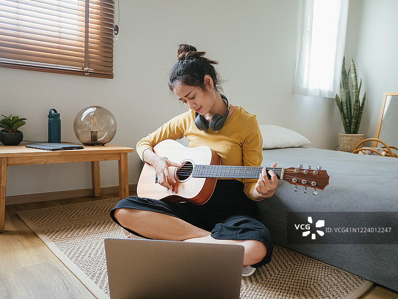 年轻女子坐在床上看笔记本电脑，手拿吉他微笑图片素材