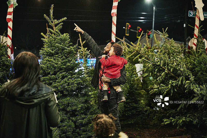 父亲抱着吃着拐杖糖的幼儿儿子，一起购买圣诞树图片素材