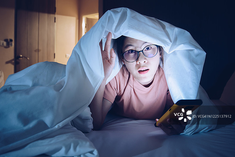深夜在床上用智能手机聊天和上网的躲在毯子下的女人肖像图片素材