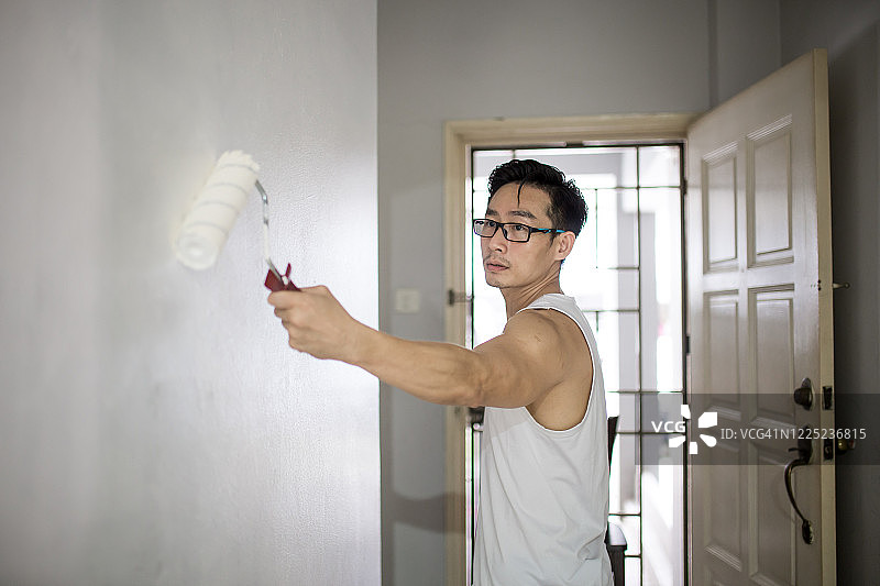 一位中国年长的男子正在用滚筒粉刷新房的墙壁，房屋改造和翻新概念。图片素材