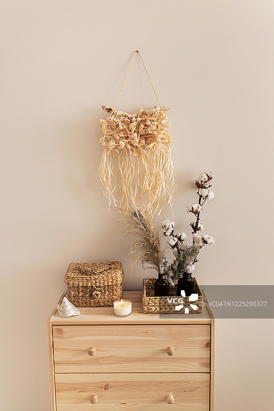 米色房间的时尚斯堪的纳维亚装饰：天然木质抽屉柜、柳条筐、干花与拉菲草编织壁挂。现代室内：米色背景墙、木制家具与手工编织壁挂，舒适家居概念图片素材