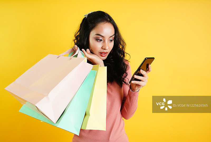 自信快乐的女人在网上购物，并通过移动应用程序订购产品。顾客的手拿着购物袋，隔离在黄色背景上。移动应用技术概念。图片素材