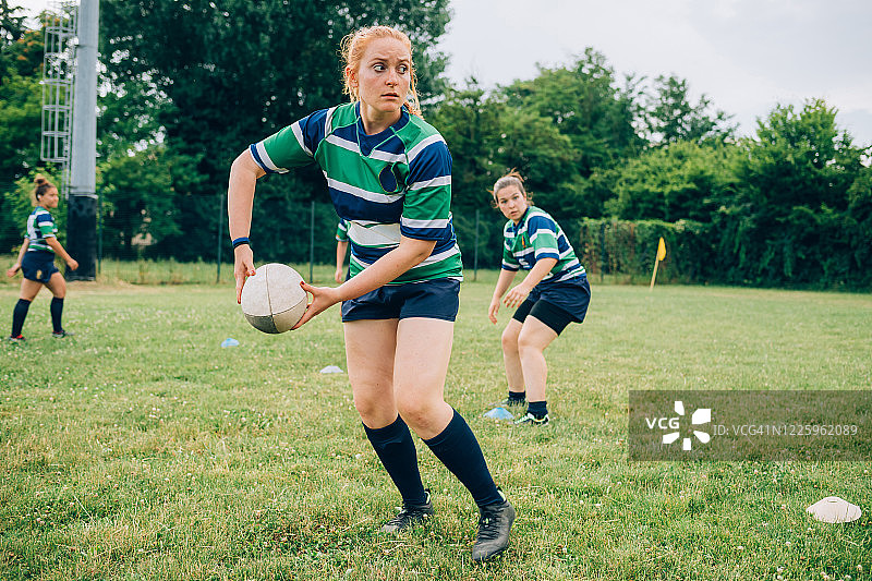 训练场上，三名女性穿着蓝、白、绿相间的橄榄球衫，其中一人准备传球图片素材
