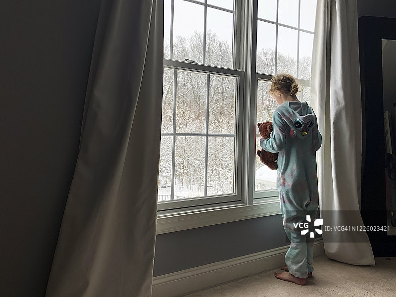 穿着睡衣连体衣的女孩抱着泰迪熊望向窗外图片素材