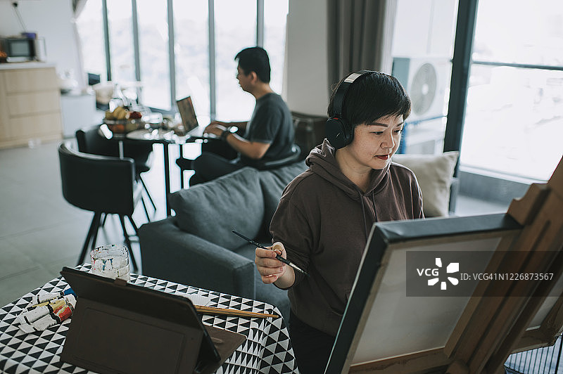 亚洲华人女艺术家在家中用调色板、画笔和画纸在画架上绘画，在客厅里戴着耳机听音乐，而她的丈夫在餐厅工作图片素材