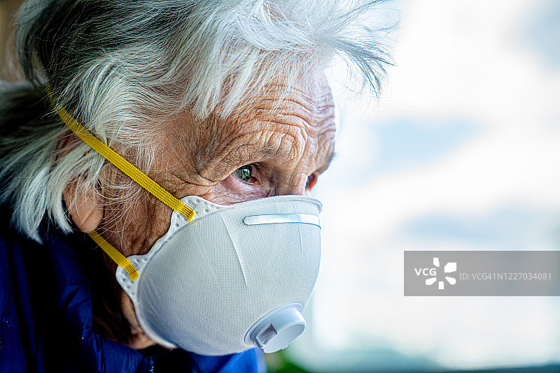 一位沮丧的老年白人妇女，戴着N95防护口罩，在寒冷和流感季节，为了防止新冠病毒H7N9流感疾病的传播，望着窗外，感到孤独的特写镜头图片素材