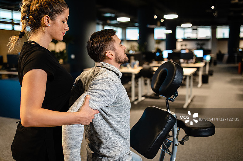 疲惫的程序员在办公室使用便携式按摩椅进行背部按摩图片素材