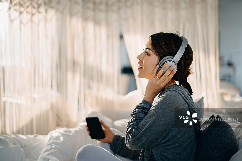 在家坐在床上，用智能手机戴耳机听音乐的放松亚洲年轻女子图片素材