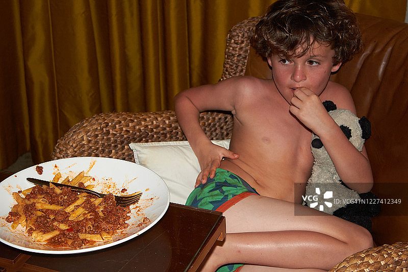 小男孩坐在舒适的椅子上，抱着熊猫玩偶，面前有一大碗意大利面图片素材