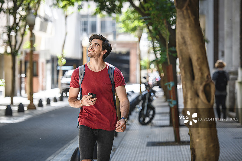 微笑的西班牙裔男游客使用智能手机走路图片素材