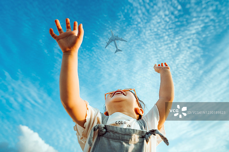 快乐的亚洲小女孩戴着花朵形太阳镜，高兴地笑着，举起手向晴朗蓝天中的飞机挥手图片素材