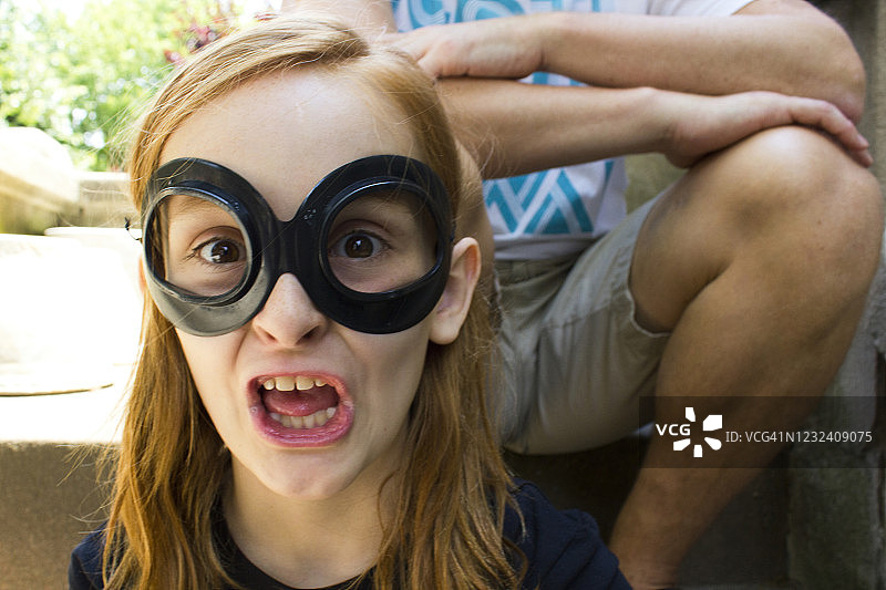 戴着面具在户外台阶上做鬼脸的红发小女孩，身后坐着一位成年人图片素材