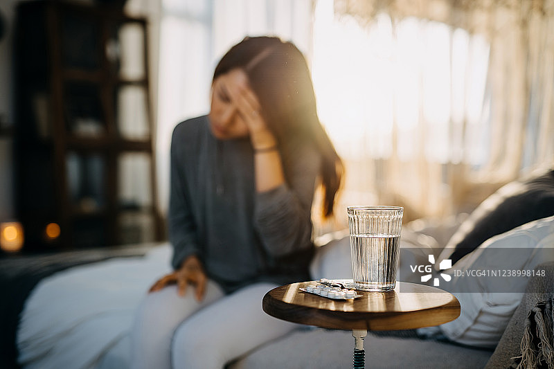 年轻的亚洲女人坐在床上感到不适，头痛难忍，床头柜上放着一杯水和药图片素材