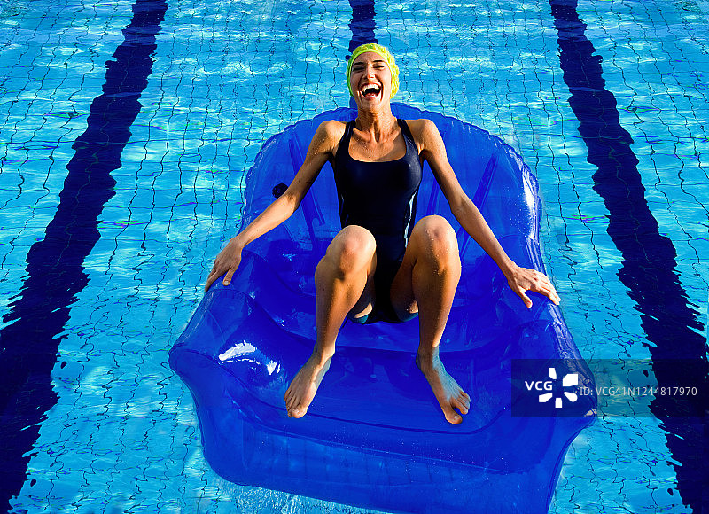 俯视：身穿黑色泳衣的女子坐在蓝色充气椅上，在户外游泳池里对着镜头大笑图片素材