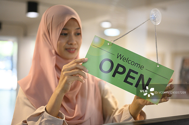 美丽的伊斯兰妇女高兴地在玻璃门上挂起“营业”标志图片素材