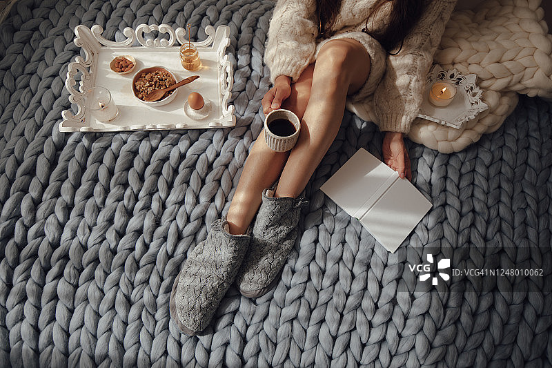 长发女人在床上喝热咖啡看书，穿着羊毛袜和毛衣，坐在厚实的羊毛格子呢上，温馨的冬日早晨图片素材