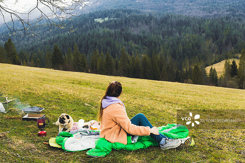 年轻女人带狗在山景附近的树旁野餐图片素材