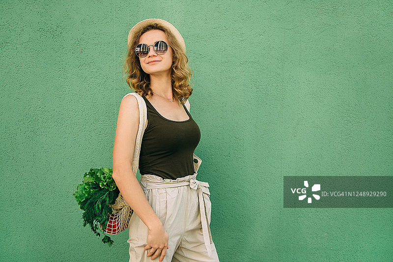 身穿浅色夏装的年轻女子，拿着装满蔬菜、绿色蔬菜和可重复使用物品的环保袋图片素材