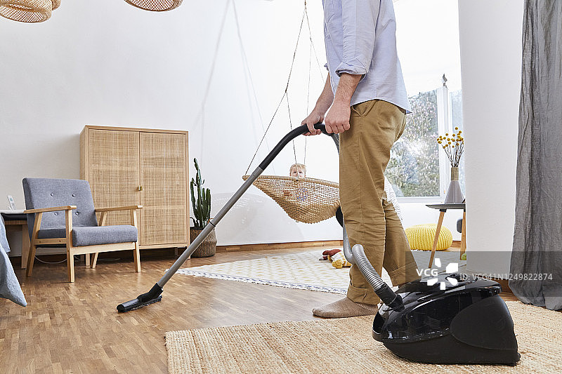 在秋千上看着父亲用吸尘器打扫客厅的男婴图片素材
