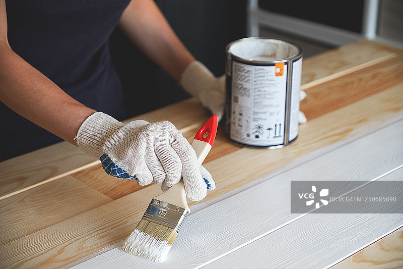 戴防护手套的女人手中的白色油漆刷。旁边是一罐油漆。绘画和修理工作。女人粉刷木板和表面，自己制作木制照片墙。图片素材