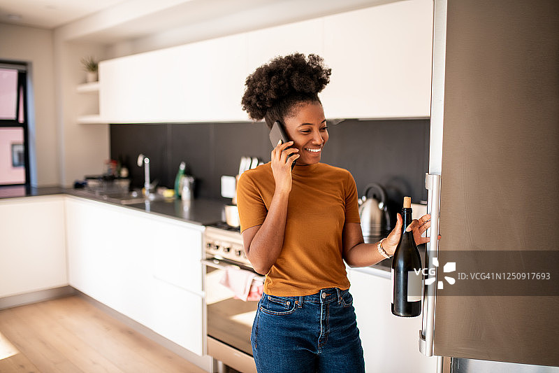 年轻女子从冰箱里拿出一瓶葡萄酒并打电话图片素材