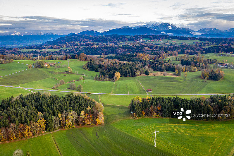 航拍视角：巴伐利亚米斯巴赫附近的秋季景观，道路延伸至远方，阿尔卑斯山脉的山峰在地平线上，多云的天空，黄色的树木和绿色的田野图片素材