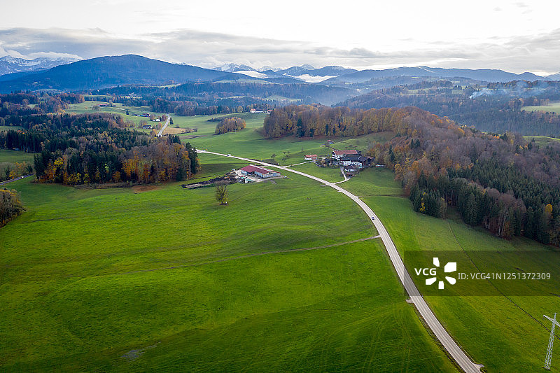 航拍视角：巴伐利亚米斯巴赫附近的秋季景观，道路延伸至远方，阿尔卑斯山脉的山峰在地平线上，多云的天空，黄色的树木和绿色的田野图片素材